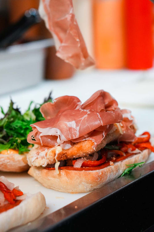 A hand-made panini with prosciutto being added onto it 